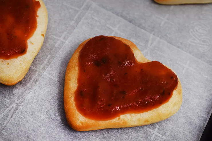 Lightly baked pizza dough, shaped like a heart, with pizza sauce spread over it.